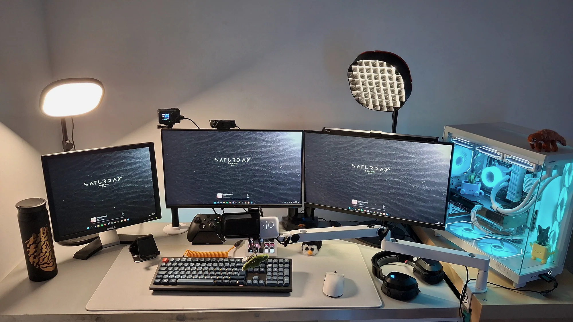 Vue d'ensemble du setup : trois écrans sur un bureau blanc, deux lumières de stream, le clavier, le Stream Deck et le PC à droite, avec ses ventilateurs éclairés en bleu clair.